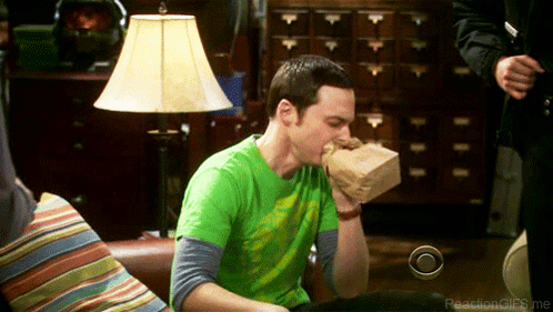 a man in a green shirt is sitting on a couch eating out of a paper bag