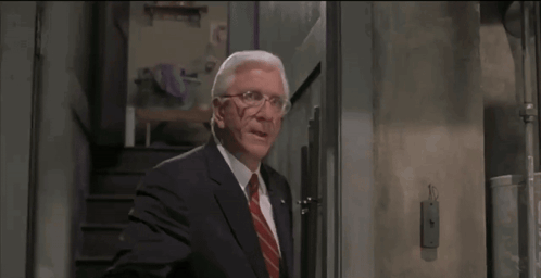 An older suited man stands in a doorway with a deadpan expression.