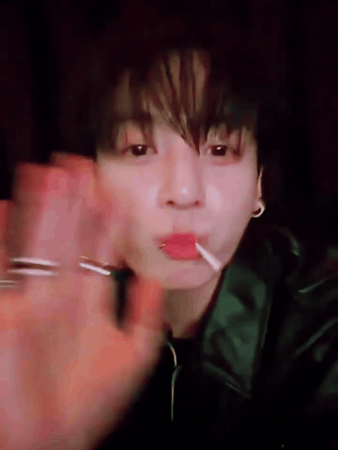 A man holds a lollipop and waves toward the camera in dim lighting.