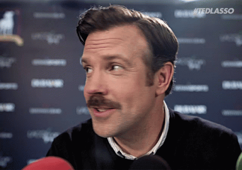 Ted Lasso glances sideways with a confident, amused smile at a press backdrop.
