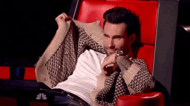 Adam Levine looks bashful while hiding behind fabric in a red chair.