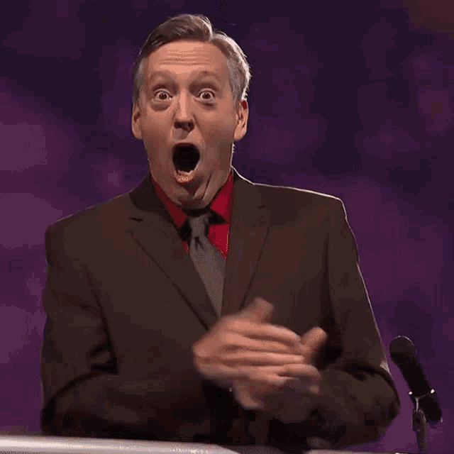 A suited game-show announcer claps with his mouth open in amazement.