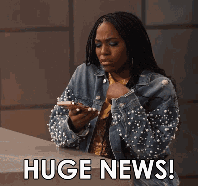 a woman wearing a denim jacket with pearls on the sleeves looks at her phone and says " huge news "