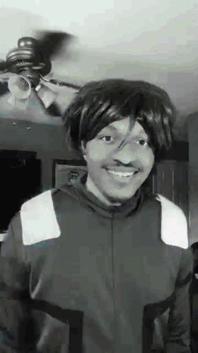 a black and white photo of a man wearing a wig and smiling in front of a ceiling fan .