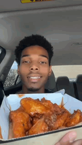 a man in a car is holding a box of chicken wings and smiling .