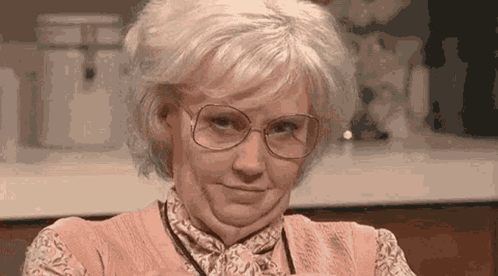 An elderly woman with glasses gives a skeptical sideways look in a kitchen.