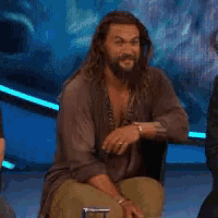 Aquaman smiles gently while seated on a studio stage.