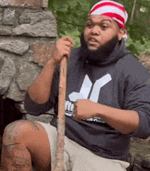 Druski holds a wooden handle outdoors while raising one hand in a shrug.