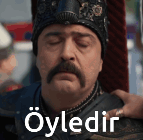 a man with a mustache is wearing a crown and says " öyledir " on his chest