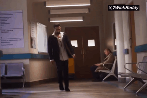 a man in a suit walks down a hallway next to a sign that says x7wickreddy