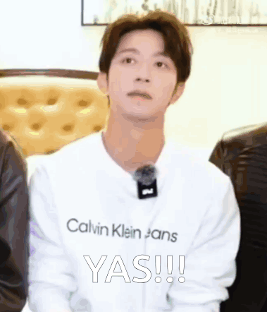 a young man wearing a white calvin klein jeans shirt is sitting in front of a group of people .