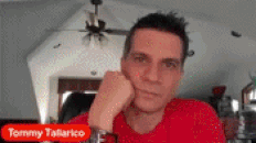 a man in a red shirt is sitting in front of a sign that says tommy tallarico