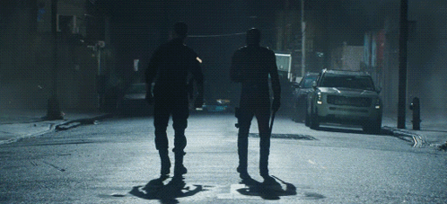 two men walking down a street at night with a car behind them