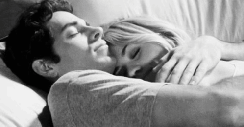A couple lies close together in bed, smiling affectionately.