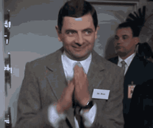Mr. Bean clasps his hands and smiles politely.