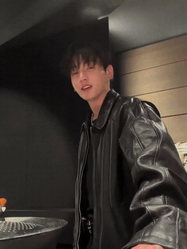 A man in a leather jacket stands in a kitchen-like room holding a lollipop.
