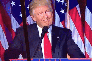 Donald Trump smiles at a podium in front of U.S. flags.