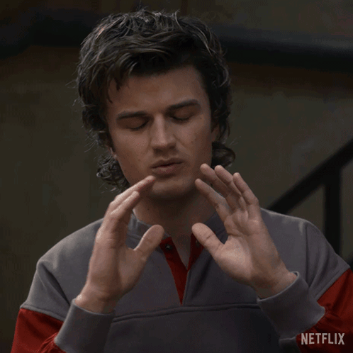 Steve Harrington raises both hands uncertainly while looking down.