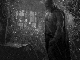 Batman sits alone on a playground swing in a rainy black-and-white scene.