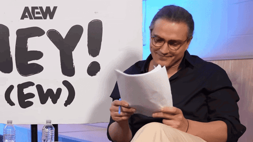 RJ City reads papers in front of a large “HEY! (EW)” sign.