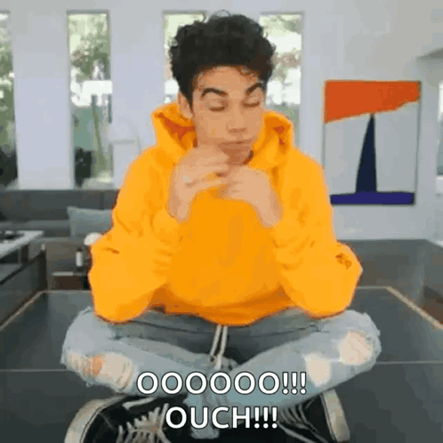 a man in a yellow hoodie is sitting on a table with his legs crossed and making a funny face .