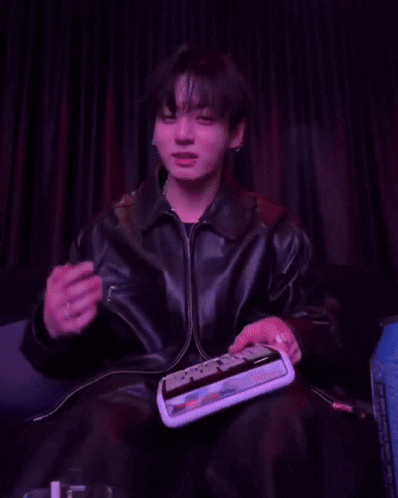 Jungkook in a dark leather jacket holds a small object against a purple-lit curtain backdrop.