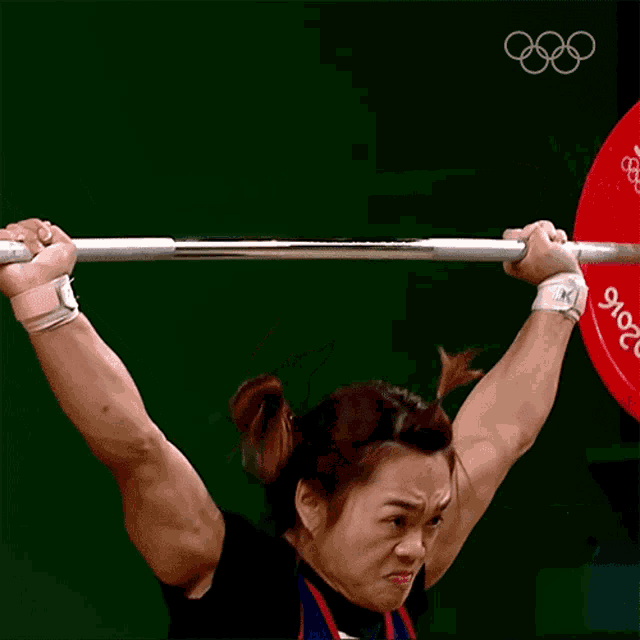 a woman is lifting a barbell with the olympics logo in the background