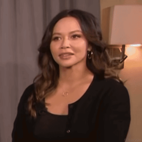 a woman in a black sweater is standing in front of a lamp and looking at the camera .