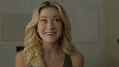 A blonde woman smiles awkwardly with an eager expression.