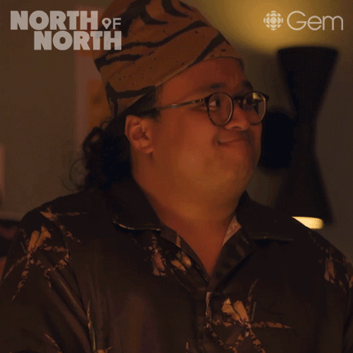 A man in glasses and a cap gives an awkward pained smile in a dim restaurant scene.