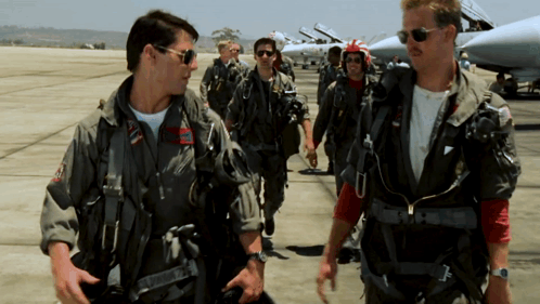 Pilots in flight suits gesture toward one another on an airfield.