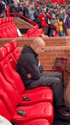 Pep Guardiola sits alone in a stadium seat looking deeply stressed.