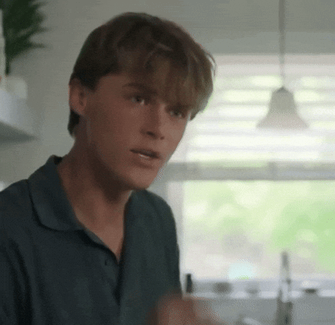 a young man in a blue shirt is standing in a kitchen talking to someone .
