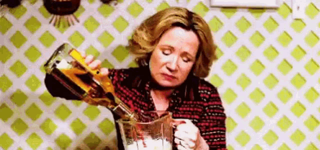 A woman pours a drink while looking dejected in a kitchen-like setting.