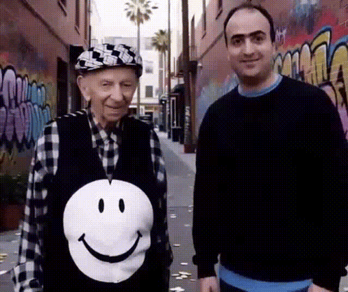 an older man wearing a smiley face vest stands next to a younger man