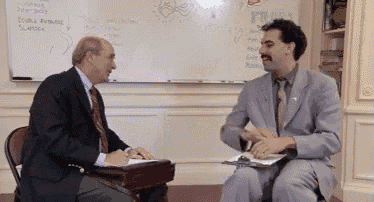 Borat sits opposite an older man and gestures during a formal conversation.