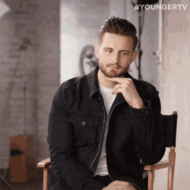 a man in a black jacket is sitting in a chair with his hand on his chin