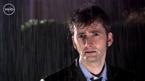 David Tennant stands soaked in heavy rain with a devastated expression.