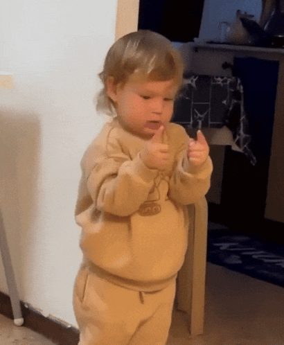 A small child gives a thumbs up while standing indoors.