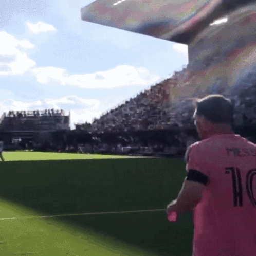 A soccer player in a pink jersey stands on a field beneath a large stadium screen.