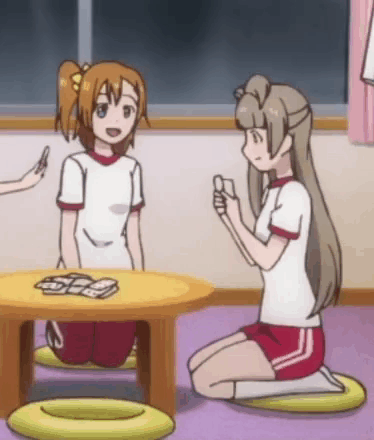 Two Love Live anime girls sit at a low table and raise hands in celebration.