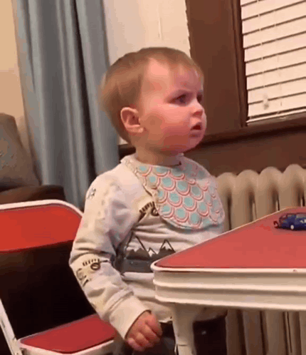 A toddler at a table looks over with a baffled expression.