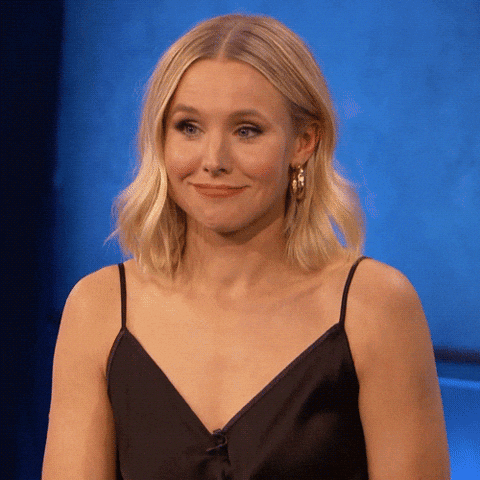 A blonde woman in a black dress gives a small uncertain smile on a blue-lit set.