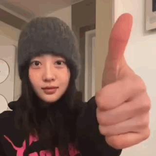 A young woman in a knit hat holds a large thumbs up close to the camera.