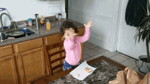 a little girl in a pink shirt is sitting at a table with her hands in the air