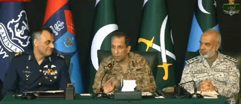 three men in military uniforms sit at a table with microphones in front of flags