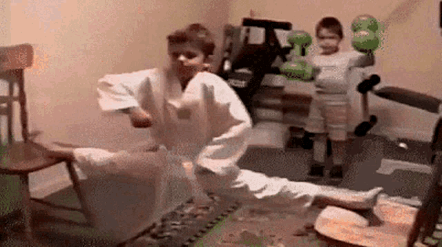 a boy in a karate uniform is kicking a chair while another boy holds a boxing glove .