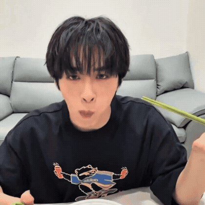 a young man wearing a black t-shirt with a bear on it is eating with chopsticks .