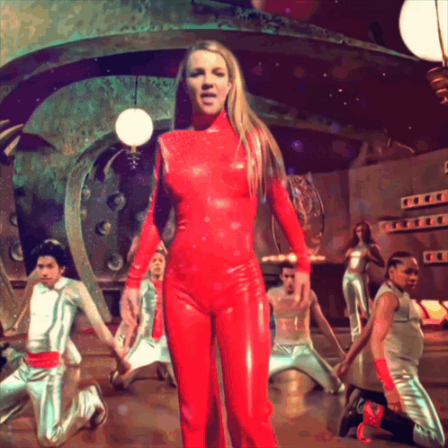 a woman in a red jumpsuit is standing in front of a group of dancers