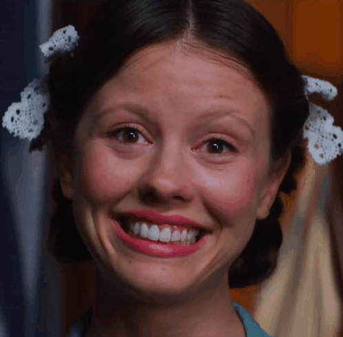 A woman with hair bows flashes an exaggerated toothy grin.